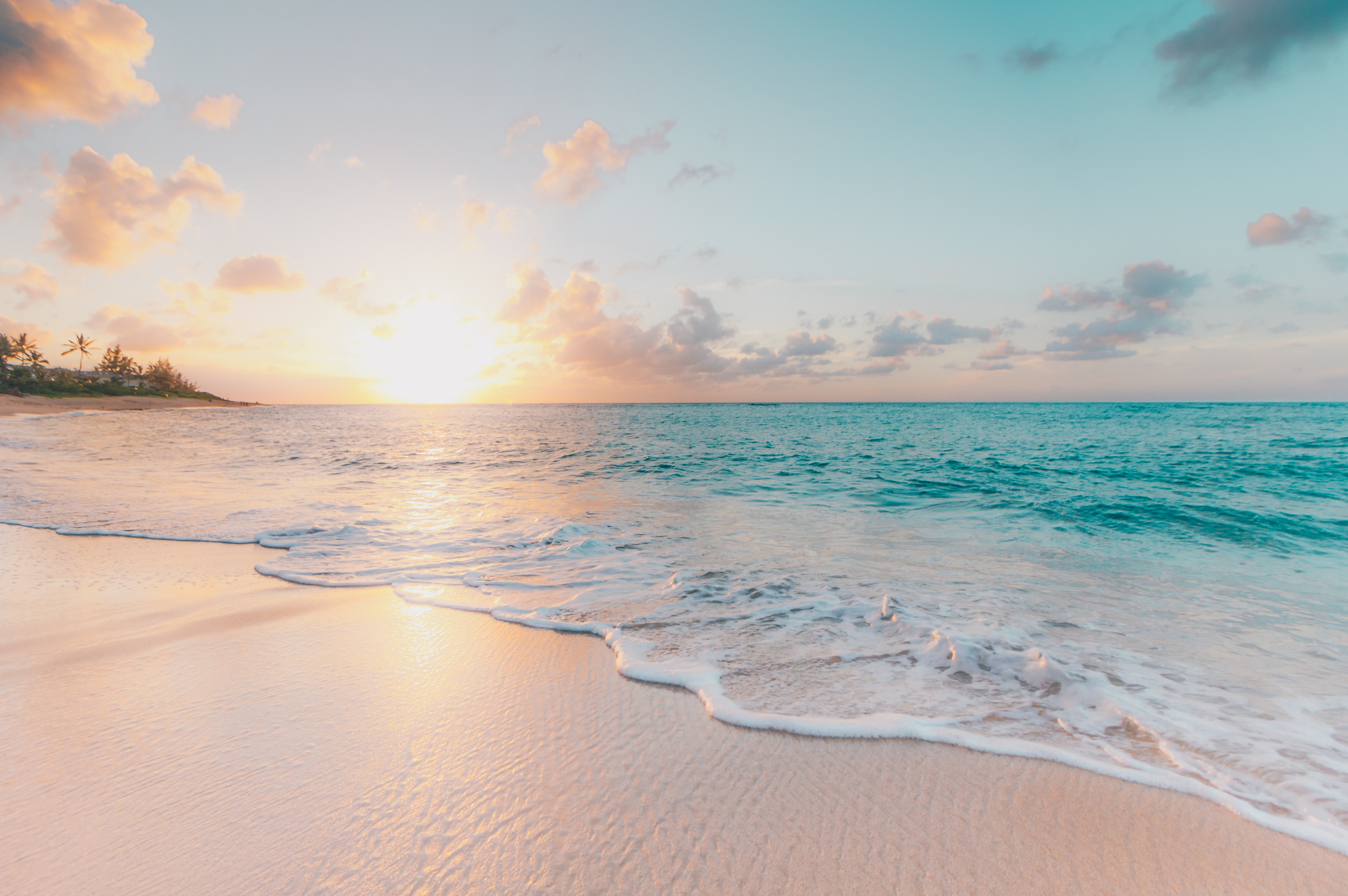 A photo of a sunset on the beach in Oahu, Hawaii. There is pale, clean sand in the foreground and blue water in the background. Photo by Sean Oulashin on Unsplash.
