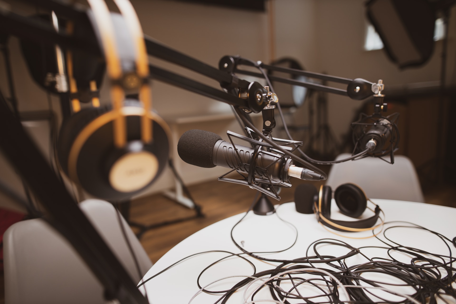 description: A podcast studio. A microphone on a boom is in focus. A set of headphones in the foreground, and the table in chairs in the background are out of focus. Photo by Jonathan Farber on Unsplash.