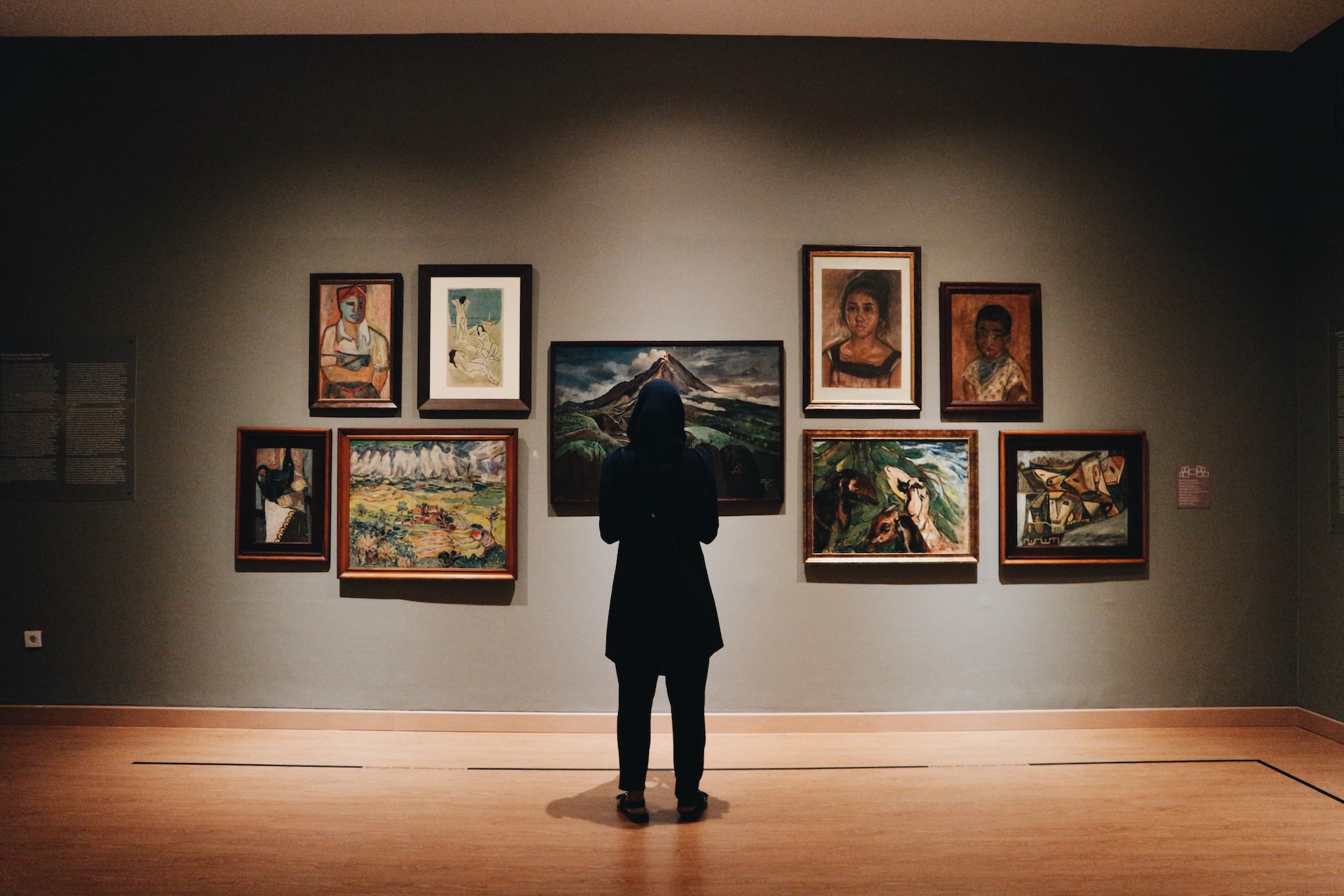 Description: A woman, in a black coat, has her back to the camera. She is facing a wall of paintings in an art gallery. Photo by Zalfa Imani on Unsplash.