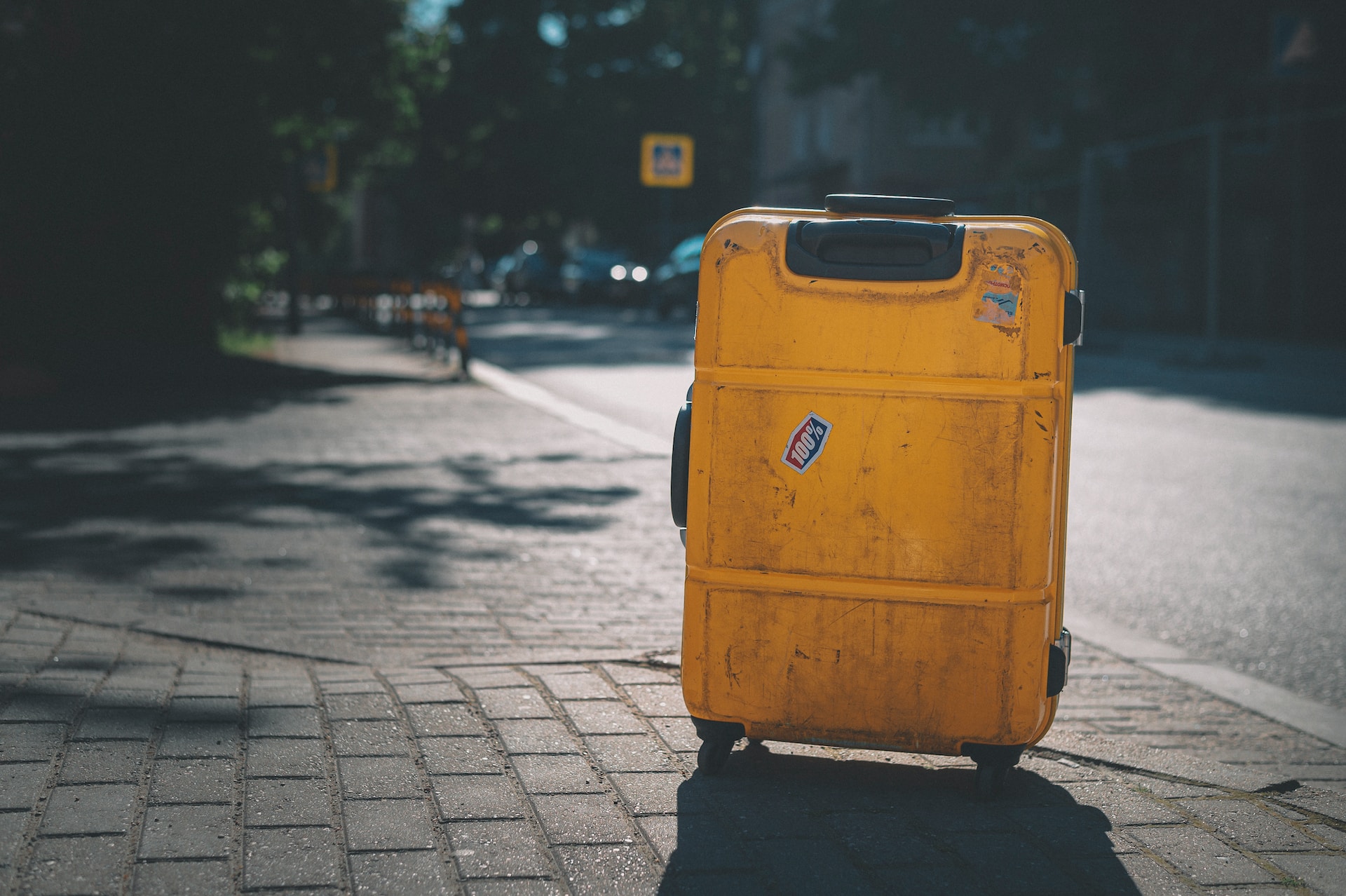 description: An image of a wheeled yellow suitcase, upright, on the sidewalk. The backdrop of a city street is slightly out of focus.