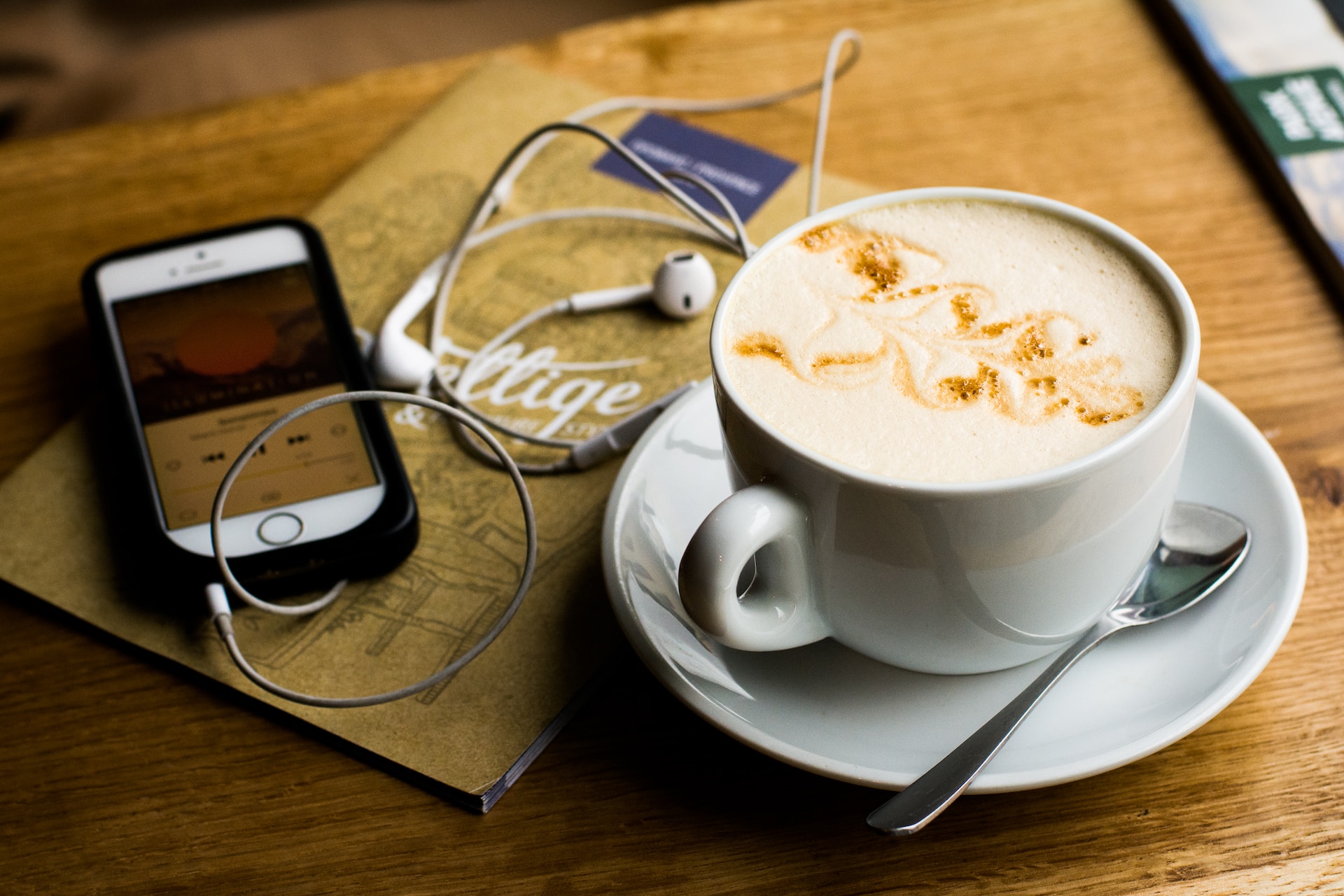 description: An iPhone and latte cup (on a saucer) sit on a wooden table. The phone has headphones plugged in. Photo by Juja Han on Unsplash