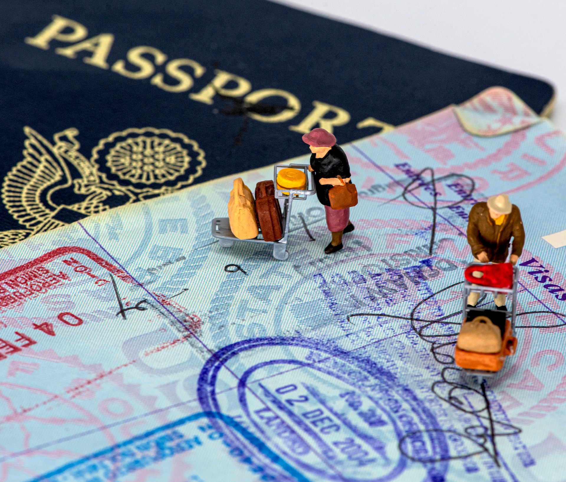 description: A close-up photo of a US passport. Two plastic figurines – people pushing luggage carts – are positioned on the page. Photo by mana5280 on Unsplash.
