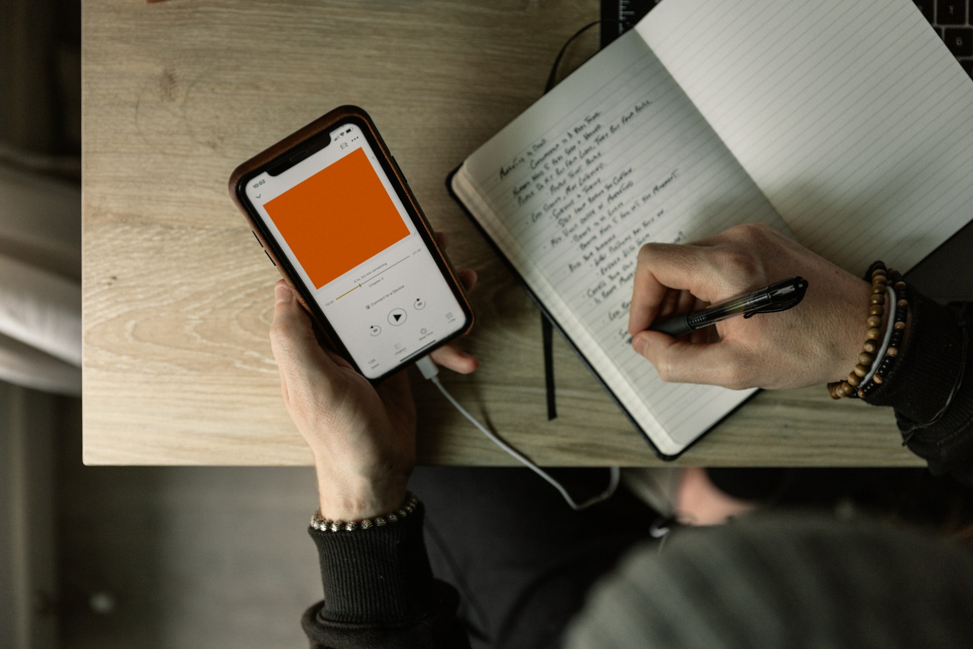 description: Overhead shot of a person, holding a phone with one hand and writing in a notebook with the other.  The phone is playing an audiobook or a podcast and it is connected to headphones.