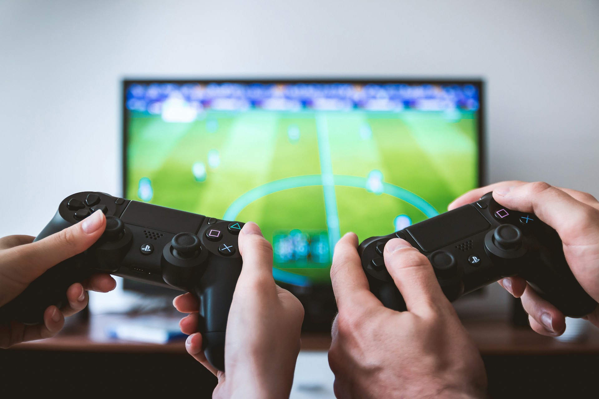 description: Two sets of hands, in the foreground, use PS4 game controllers.  In the background, which is out of focus, a TV shows the video game in play.  Photo by JESHOOTS.COM on Unsplash.