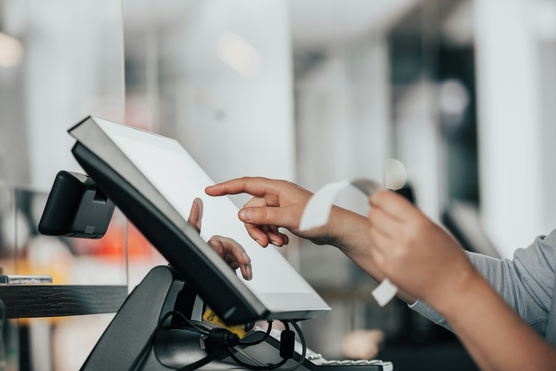 description: A close-up shot of a salesperson working a touchscreen cash register.  One hand is tapping the screen while the other is holding a receipt.  Photo by Simon Kadula on Unsplash.