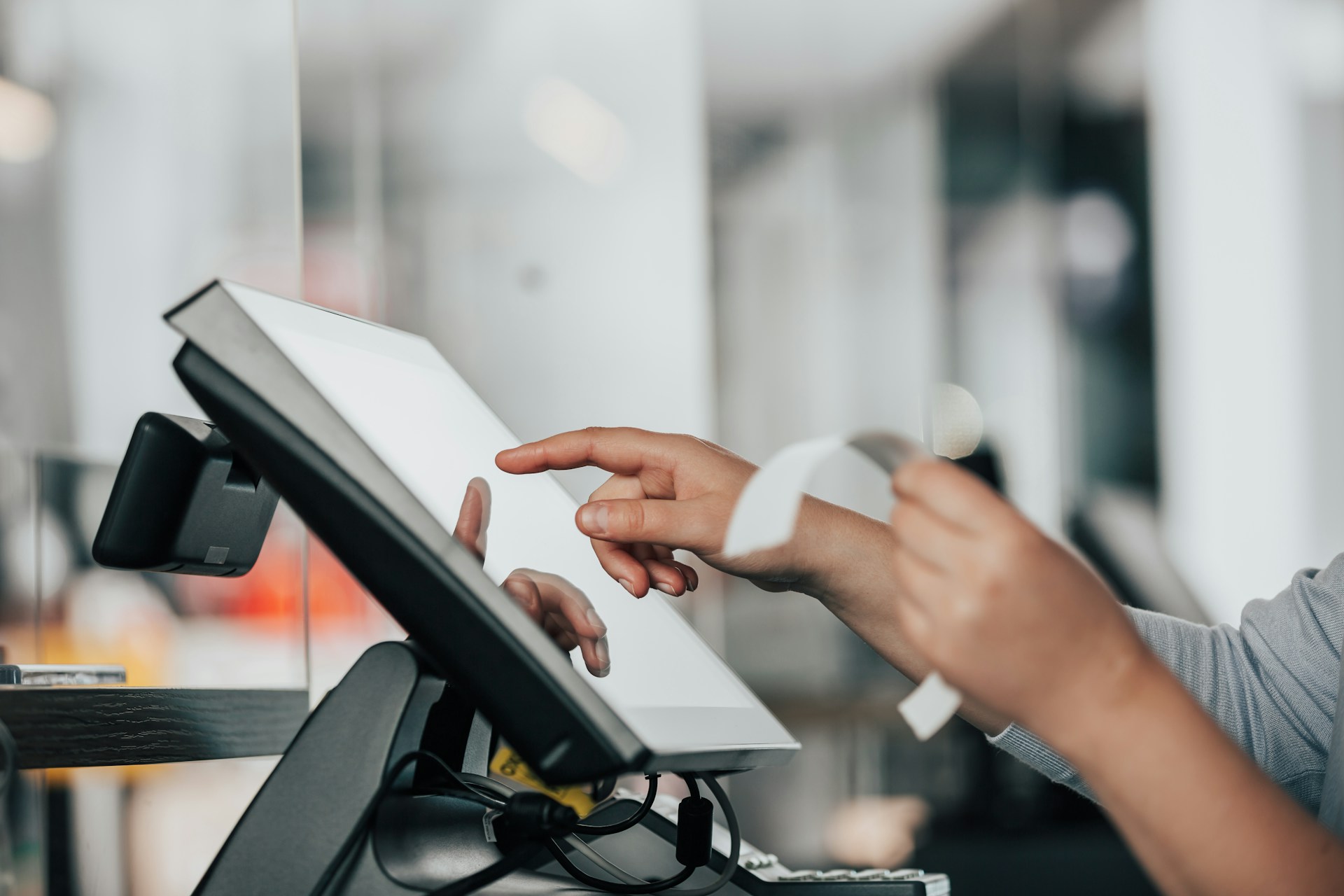 description: A close-up shot of a salesperson working a touchscreen cash register. One hand is tapping the screen while the other is holding a receipt. Photo by Simon Kadula on Unsplash.
