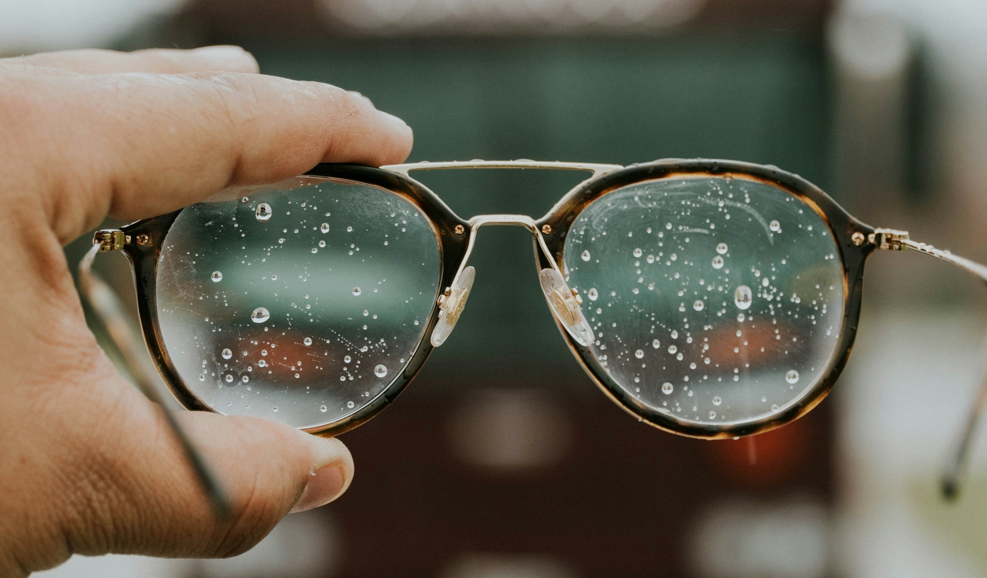 description: A hand holding up a pair of glasses. The lenses are streaked with droplets, making it difficult to see through them. Photo by Hermes Rivera on Unsplash.